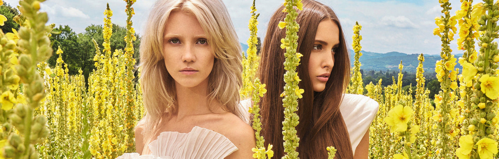 Zwei junge Frauen in einem gelben Blumenfeld. Eine mit blondem, voluminösem Haar und eine hat braunes, glattes Haar. Alles wirkt sehr natürlich.